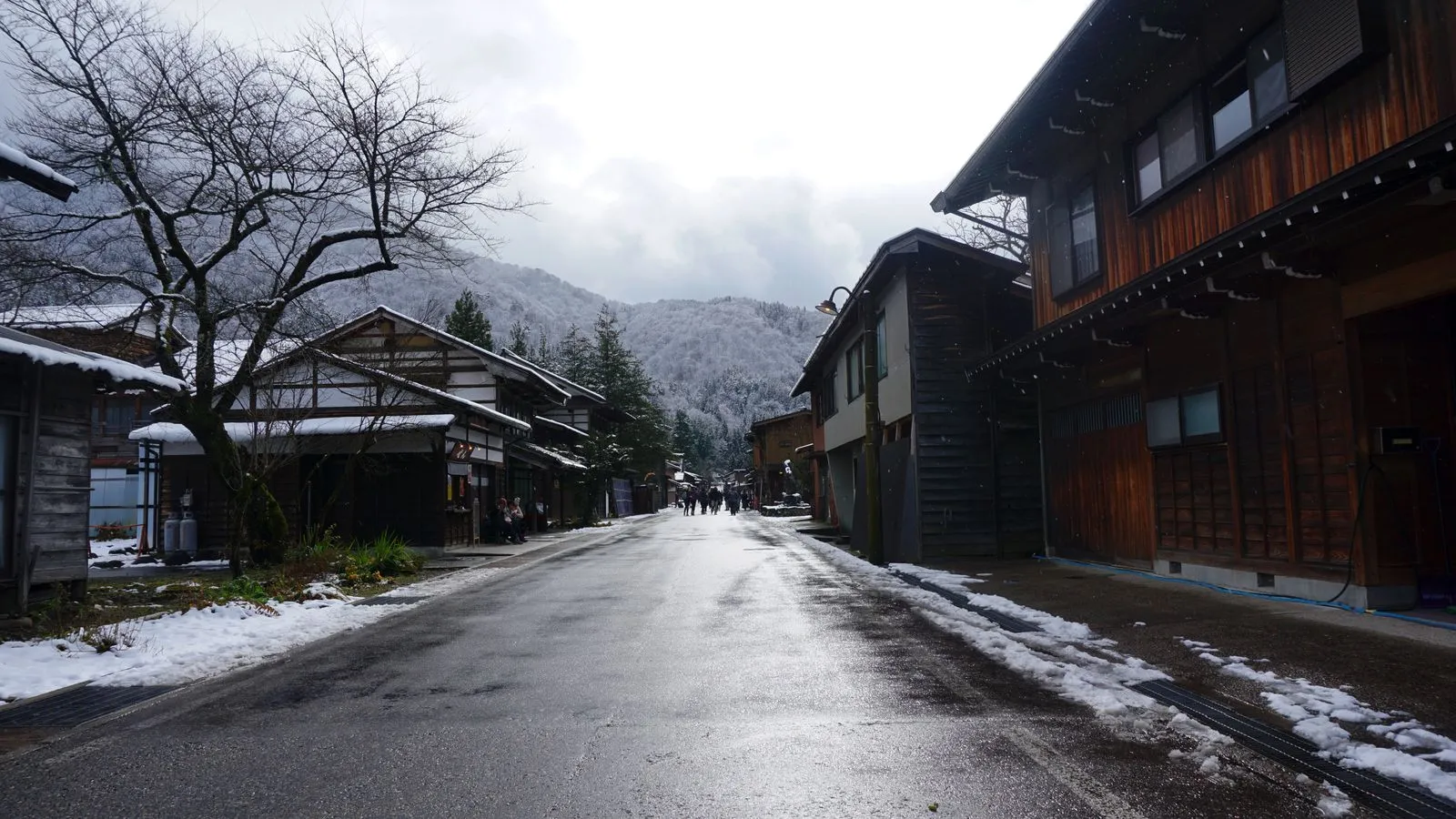 Wet road through Shirakawa-go with dark timber houses and snow on the hills