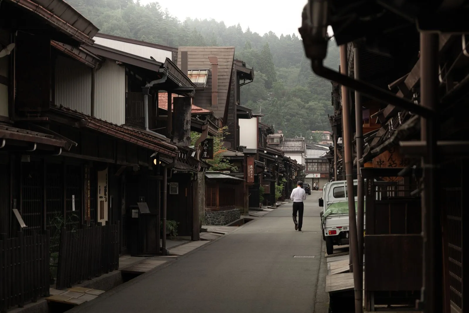 A quiet lane of dark wooden houses with deep eaves, a forested hill behind and one person walking away up the street