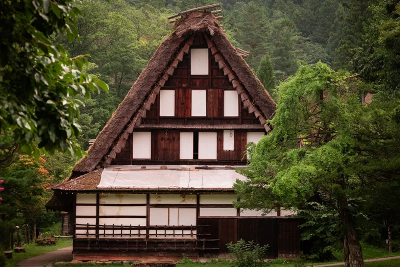 Thatched gassho-zukuri farmhouse with white paper windows among trees at Hida Folk Village