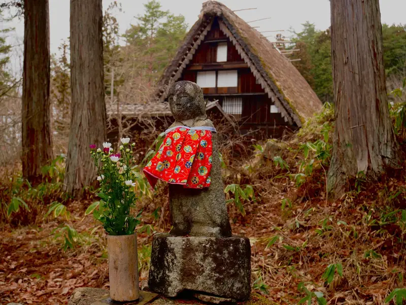 Stone jizo in a red bib with flowers in front of a thatched farmhouse at Hida Folk Village