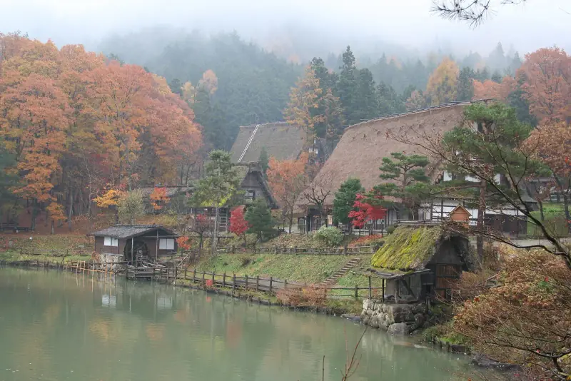 Thatched farmhouses and a water mill around the pond at Hida Folk Village in misty autumn colour