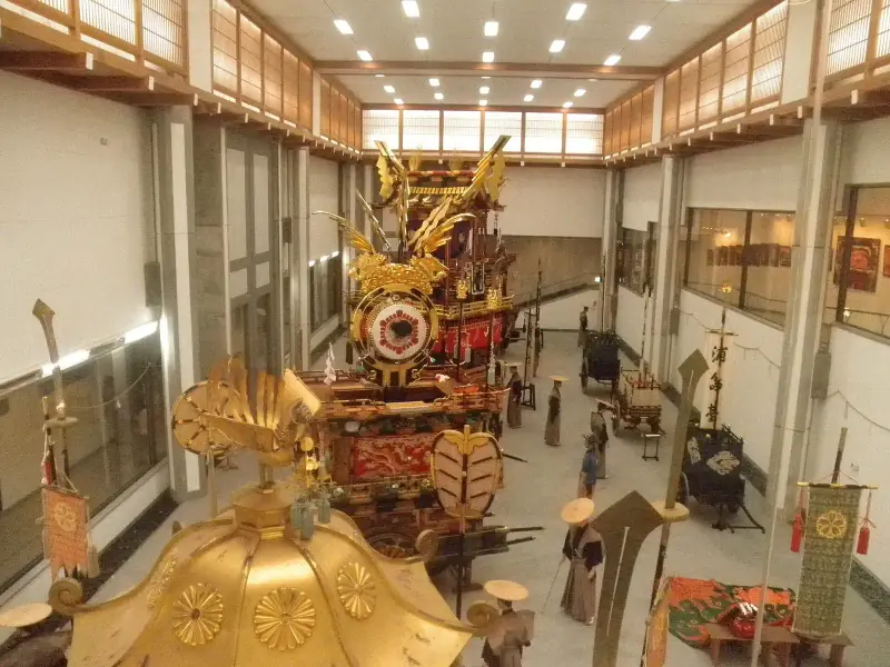 The Takayama Yatai Kaikan hall from above: a gold mikoshi in front, a decorated festival float and costumed figures behind
