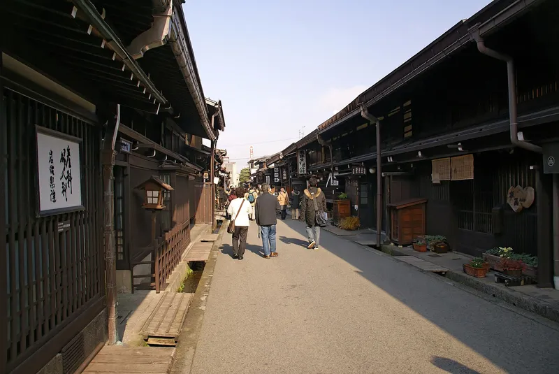 Visitors strolling a sunlit Sanmachi street of dark merchant houses with water channels