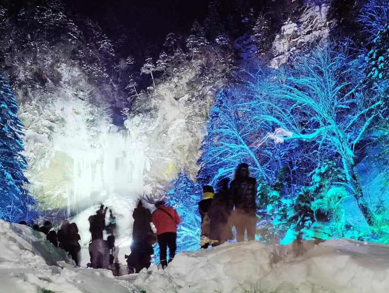 Frozen Hirayu Falls lit up at night in winter, snow-laden trees glowing blue, visitors seen from behind