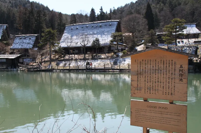 Goami pond at Hida Folk Village with snow-dusted thatched roofs and its information board